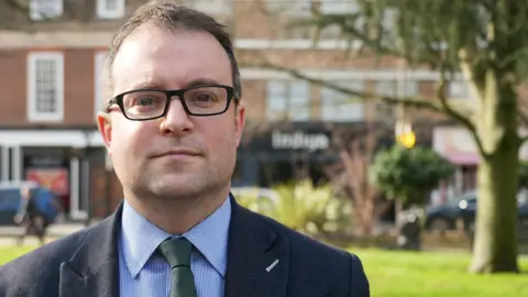 Ant Saddington/BBC Lib Dem opposition leader Henry Vann is standing in a grassy outdoor area with trees, shrubs, and open space behind them. Further back, across the street, there are brick buildings with large windows and storefronts, including several shop signs. A few cars and pedestrians are visible in the background. The person is wearing a dark textured jacket over a blue check-patterned shirt and a green tie.