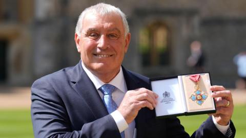 Peter Shilton wearing a blue suit and holding up his CBE in a box at Windsor Castle