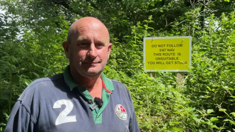 BBC Mark Nicholls wearing a grey T-shirt stood in front of a hedge. In the hedge is one of his signs which reads: "Do not follow sat-nav this route is unsuitable you will get stuck."