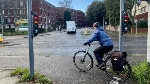 A cyclist in a light blue jacket and dark trousers with a pannier at cycling traffic lights on Western Way looking up towards Barnfield Road