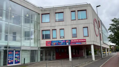 A glass-fronted school building extends towards some tower blocks on a grey day, as seen from across a roadway. 