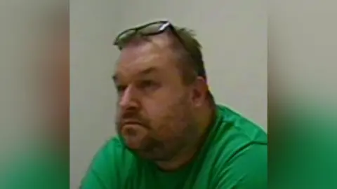 A man with grey hair and stubble is wearing a green t shirt and his glasses are on top of his head. He is sitting in front of a white wall.