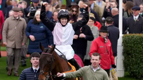 Adam Davy/PA Rachel Blackmore is smiling with her arms up sitting on a brown horse. She is wearing a black white and light pink silk with a black helmet decorated with a pink star.