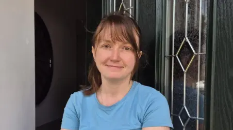 Michaela smiles as she poses outside her front door. She is wearing a blue top and has shoulder-length brown hair.