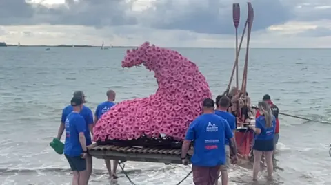 People are lifting a raft into the waves. The raft holds a sculpture in the shape of a wave which is covered in pink gerbera flowers. There is a group of make and female rowers holding the raft and others in s boat holding the oars aloft. There are yachts sailing on the horizon.