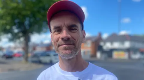 A man with a red cap stands by a road. He has a short grey beard and wears a white t-shirt. He is standing on a street corner and there are buildings and cars behind him. 