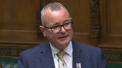 House of Commons Lee Barron wearing a navy blue blazer, white shirt and yellow and black tie speaking in the House of Commons. 