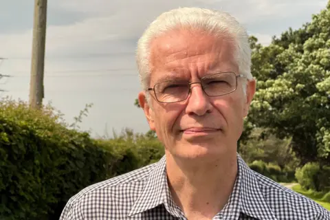 David Mudge is wearing glasses and a check patterned shirt, and has short, white hair. He is clean shaved. There are trees and hedgerows in the background.