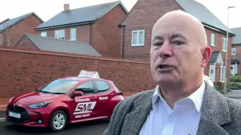 Stephen Lambert is bald and wears a grey jacket over a white, open-necked shirt. He stands in the street near his driving school car.