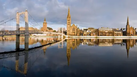 Historic buildings on Inverness' riverside, including churches, reflected in the mirror-like surface of the River Ness on a bright but cloudy day.