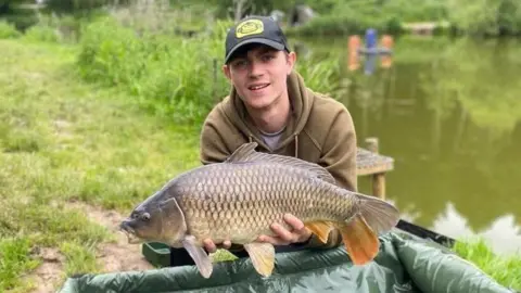 A man wearing a black baseball cap and brown hoodie is crouched on a patch of grass close to a large area of water. He is holding a large fish.