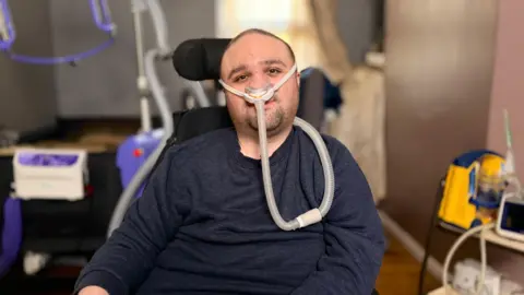 Man in his thirties with short dark hair and stubble, wearing a dark blue jumper looks at camera. He is sitting in a powered wheelchair with a ventilation tube attached to his nose. In the background is medical and hoisting equipment.