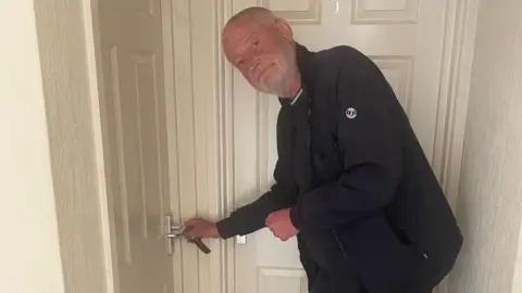 Joe is turning a key in a door to a home. He is wearing all black and looking at the camera and smiling. The door is white and another door joins it on the right.