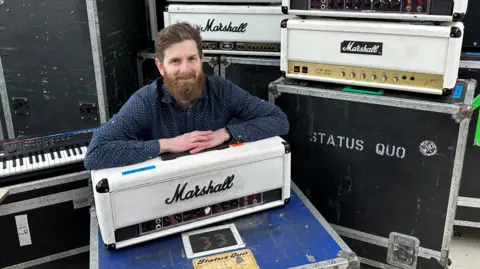 Image show Luke Hobbs surrounded by Status Quo band equipment and leaning on a white Marshall amplifier 