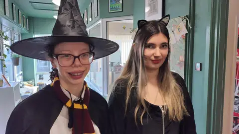 James Grant/BBC A girl in a large black witches hat, glasses, a maroon and yellow scarf, and black cloak stands next to a girl in a black dress wearing cat ears and whiskers painted on her cheeks. Both are smiling at the camera. 