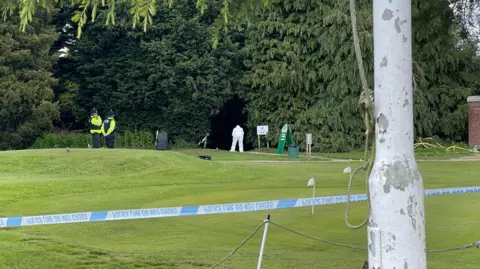 Police tape at the golf course behind a tree. Figures can be seen in the distance around a path.
