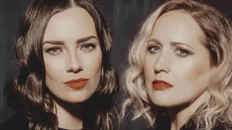 Kris Kesiak Marissa Keltie, who has dark hair, and Caroline Gilmour, who has blonde hair, look directly into the camera.