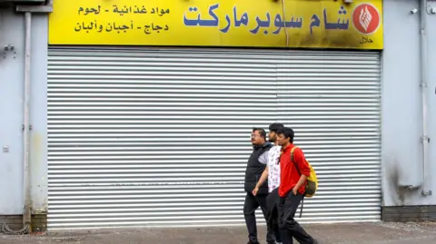 Getty Images People walk past a Syrian supermarket in Belfast city centre on August 10, 2024, which was attacked by anti migrant protesters. The three people are men, one in a red top wearing a yellow bag pack, one in a white top and one in a black top. All three have black hair. The supermarkets shutter is down, the storefront above the shutter is yellow and shows signs of being burned.