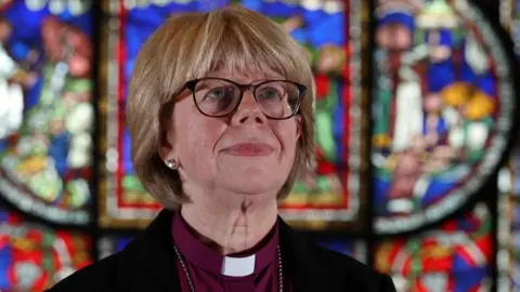 Dame Sarah Mullally, wearing a burgundy top, back jacket and white clerical collar, stood in front of a stained glass window.