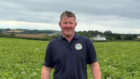 Richard Orr standing in front of a potato field. He has short dark hair, wearing a navy polo shirt.