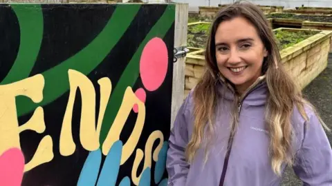 A woman with long fair hair and blonde highlights smiles into the camera. She is wearing a purple coat. She is standing beside a hand painted multicoloured sign that says 'Friends of the Field'. She is standing in front of wooden planters with green plants inside.