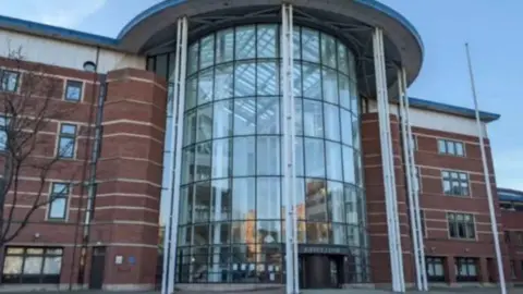 A tall brick building with a glass-fronted tower to the front. The building is set against a sunny blue sky.
