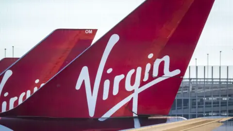 Getty Images Two planes on the ground with the Virgin Atlantic logo on their tails. The colours are red and white. A fence is to the right. 