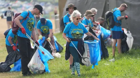 PA Media People dressed in blue T-shirts are picking up litter in the grass on the festival site.