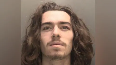 Merseyside Police A mugshot of Connor Chapman who has long curly hair, a brown moustache and wispy stubble, and smirks at the camera 