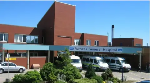 University Hospitals of Morecambe Bay NHS Foundation Trust Furness General Hospital is a red brick building with blue and white signs that read: "NHS Furness General Hospital". Ambulances are parked out front.