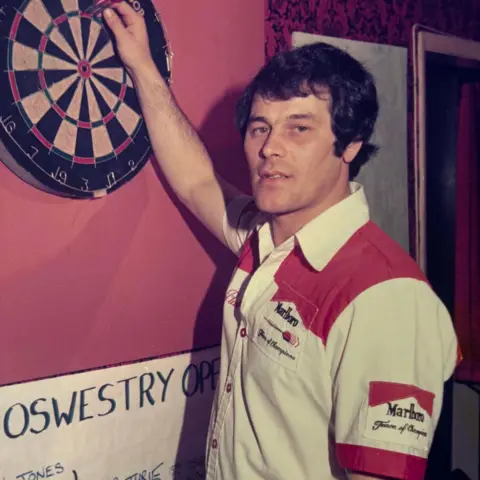 Getty Images Rab Smith stands at a dart board with a white shirt with red shoulders. He has dark hair and is picking his darts out of the board