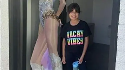 Handout A smiling Alice stands outside the Hart Space dance studio next to a life-size cardboard cut-out of Taylor Swift, wearing a black t-shirt with the slogan 'Vacay Vibes' written in colourful letters. 