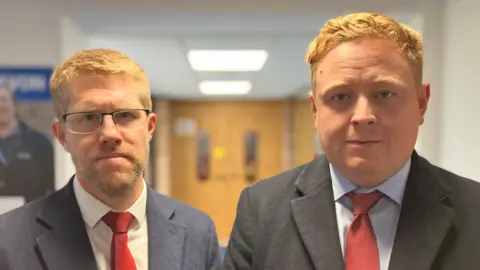 Tom Collins and Chris Bloore, both wearing suits with red ties, stand in a corridor with a pair of double doors in the distance behind them.