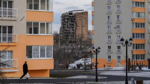 EPA-EFE/Rex/Shutterstock Um bloco danificado de apartamentos atrás de dois novos blocos construídos pelas forças ocupantes da Rússia em fevereiro de 2025