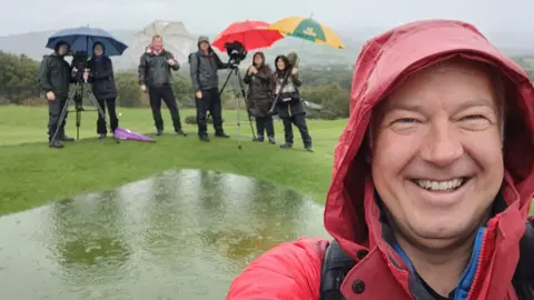 Derek Brockway BBC meterologist smiling in the rain, with TV crew from his programme Weatherman Walking