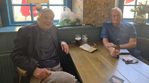 Hayley Coyle/BBC Two people sit at a wooden table in a pub beside a window. One person wears a dark jacket and shirt, while the other wears a blue short-sleeved top and holds a bottled drink. On the table are two glasses, an open notebook, a smartphone, a pair of glasses and a wallet-like item. The interior features blue-painted wood panelling, textured walls and natural light coming through the windows.