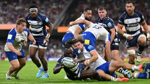 Bath Rugby and Bristol Bears players in a tackle. There are eight male players in the shot. One Bristol player has fallen to the ground and is holding the ball, while his opponent from Bath is trying to grab it from him. The crowd can be seen in the distance. 