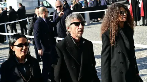 Stefano RELLANDINI / AFP via Getty Images A group of mourners dressed in black walk together outside the church after the service