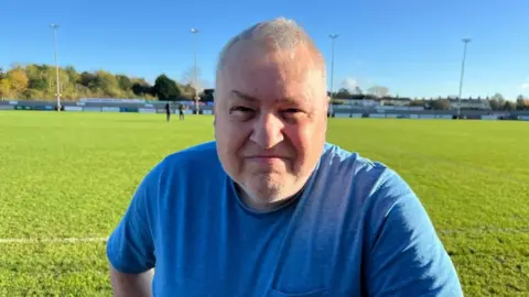 Roger Rose in a blue T-shirt smiling, while stood on the edge of a football pitch.