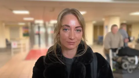 Kate Fisher is smiling at the camera as she stands in the airport. She has long blonde hair and is wearing a black furry jacket and has a handbag looped over her shoulder. Blurred behind her are two passengers pushing trolleys.