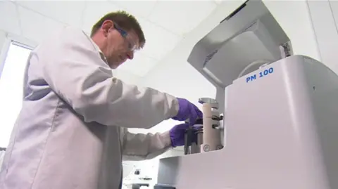 A man wearing safety goggles, a white lab coat, and purple nitrile gloves handles a piece of scientific equipment