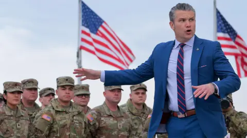 Pete Hegseth gestures in front of troops standing in front of American flags