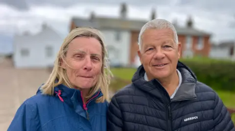 Martin Giles/BBC Lorraine and Paul Harrison are wearing blue coats and look windswept. Lorraine's coat has a pink zip and Paul's has a logo which says "Karrimor". They are standing on a path with cottages in the background. 