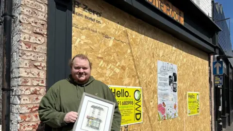 BBC A man in a green hoodie stands in front of a boarded up shop front in the sun. He is holding a framed drawing of what his shop looked like before the crash.