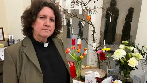 A church curate, wearing an olive blazer, a black top and a dog collar, looks at the camera as she stands in front of a prayer tree, flowers and cards. She has shoulder length brown curly hair.