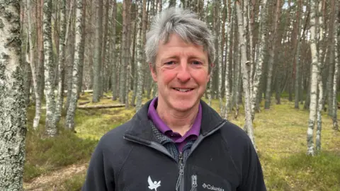 Colin Simpson is standing in an area of natural woodland. He has thick grey hair and is wearing a black fleece over a dark grey top and purple polo shirt. 