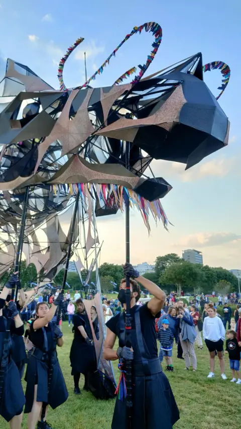 Victoria Winward Puppeteers dressed in black hold a skeletal looking dragon puppet on sticks while crowds watch on. It takes several people to manipulate the structure which is the size of a car.