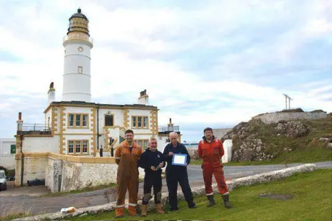 Ross Russell Ross, two engineers and the lighthouse keeper stand outside Corsewall Lighthouse. They are wearing workmen jumpsuits. The lighthouse is white.