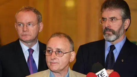 PA Media Martin McGuinness, Denis Donaldson and Gerry Adams are all looking to their right. All three are wearing glasses. Mr McGuinness and Mr Adams are both wearing dark suits, shirts and ties. Mr Donaldson is wearing a tanned coloured jacket and a denim shirt. There are three microphones in front of them.