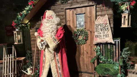 Someone dressed as Santa at night time standing outside a wooden hut decorated with festive green foliage, lanterns, Santa's canvas bag and a small child's sledge propped at the side